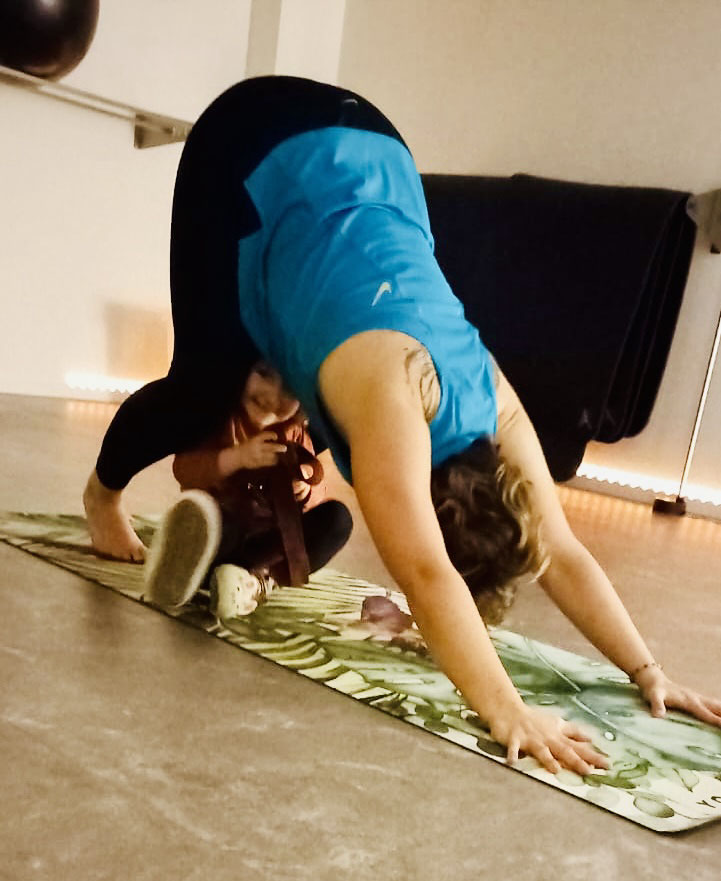 Sabine im herabschauenden Hund auf der Yogamatte, ein Kleinkind spielt unter ihr