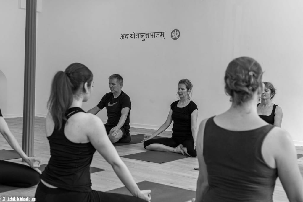 Gruppe im Sitzen im athayoga Studio, an der Wand der Sanskrit-Vers atha yoga anushasanam