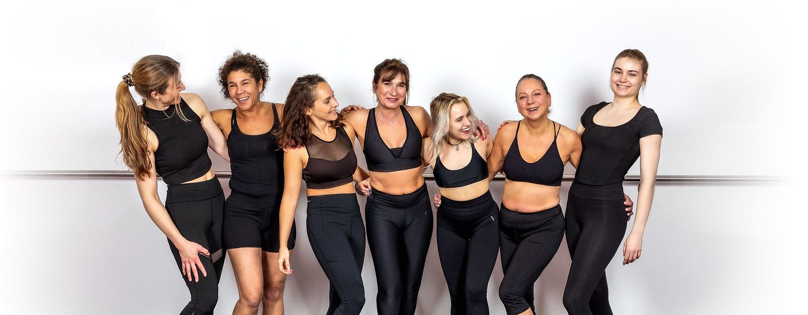 Sieben lachende Frauen des Body-&-ART-Teams Arm in Arm vor heller Wand