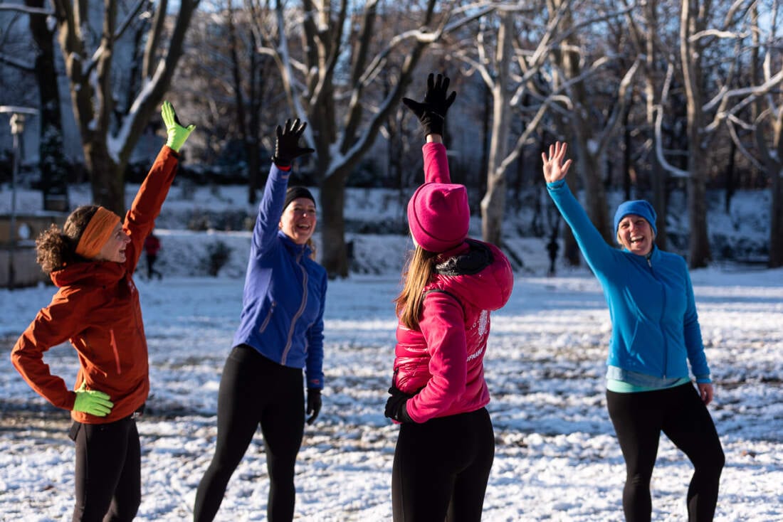 Vier Frauen in Winterjacken strecken lachend die Arme in die Höhe, um sie herum verschneiter Park