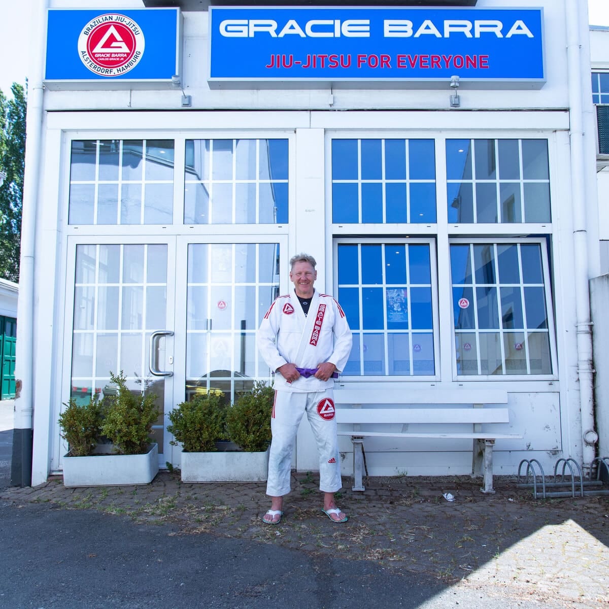 Christian Schön im weißen Gi vor dem Eingang von Gracie Barra Alsterdorf mit dem Schriftzug Jiu-Jitsu for everyone