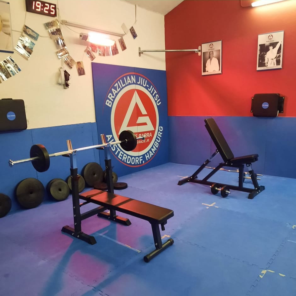 Der Kraftraum von Gracie Barra Alsterdorf mit Hantelbank und dem großen Logo auf der Mattenwand