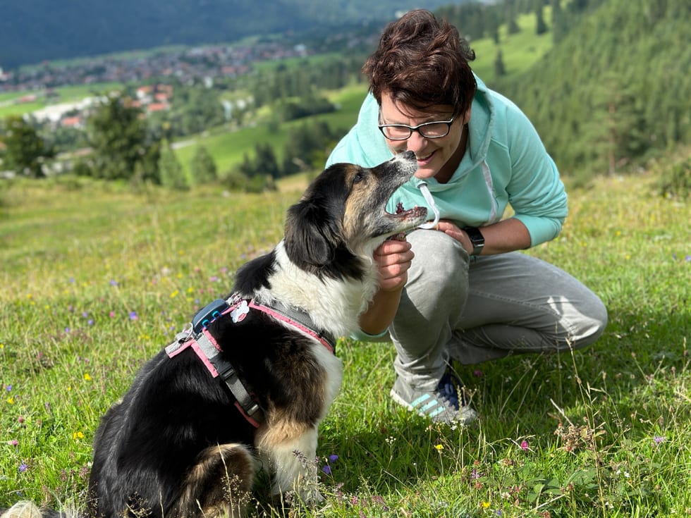 Andrea Wiesböck hockt auf einer Bergwiese über dem Tal, ihre Hündin Neela schleckt ihr übers Kinn