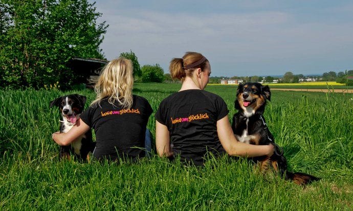 Susanne und Maruschka von hundumglücklich Maintal sitzen mit zwei Hunden auf einer Wiese, auf den Shirts das Logo