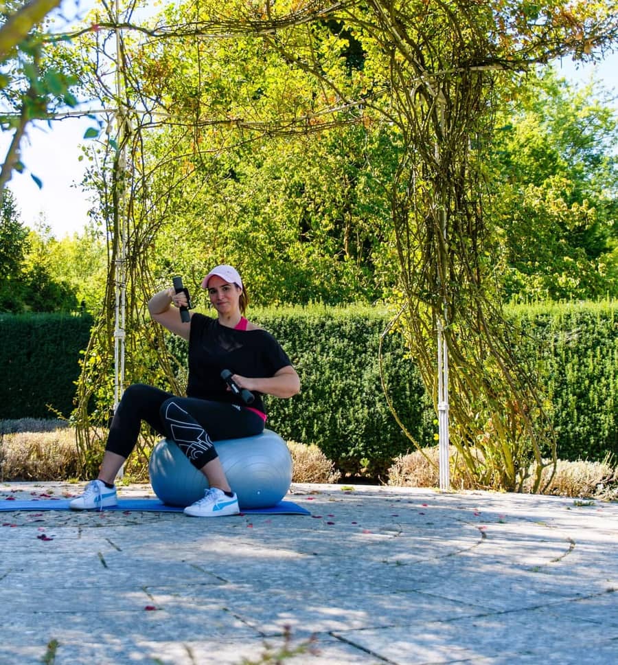 Julia sitzt beim Training mit Kurzhanteln auf dem Gymnastikball unter einem begrünten Rosenbogen im Park
