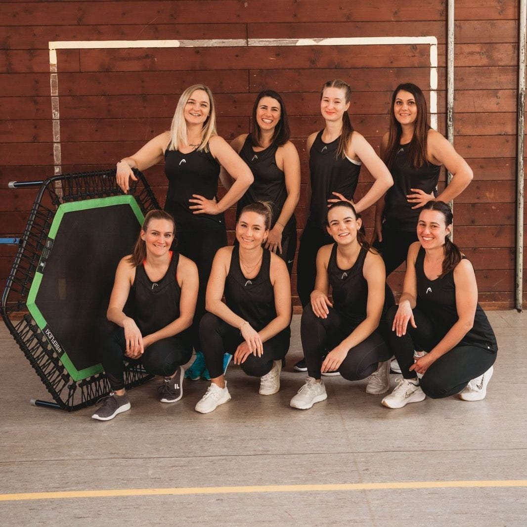 Acht Frauen in schwarzen Shirts posieren in einer Turnhalle neben einem Jumping-Trampolin mit grüner Kante