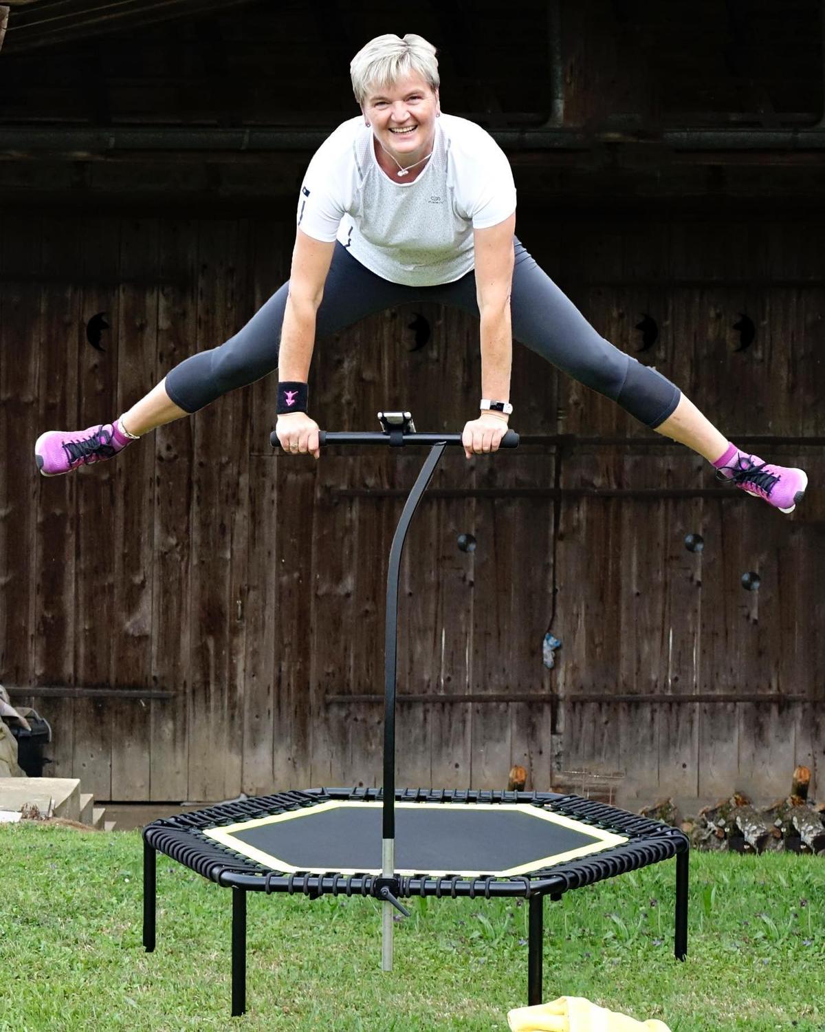 Bojana springt lachend mit gegrätschten Beinen über ihrem Trampolin, vor einer alten Holzscheune