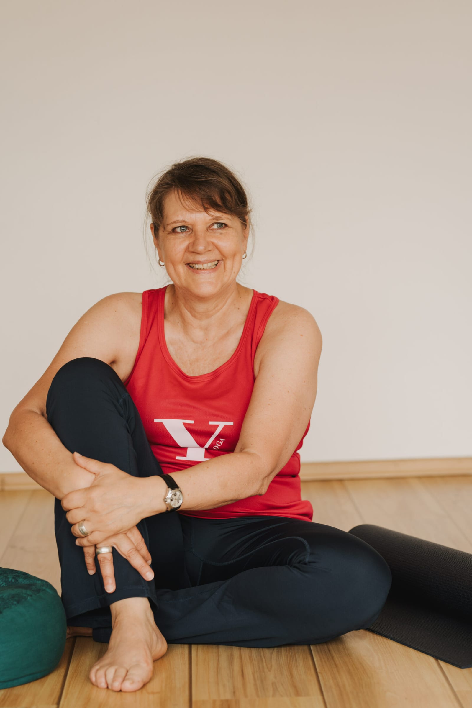 Manuela Förg sitzt lächelnd auf dem Holzboden ihres Yogaraums, neben ihr eine gerollte Matte