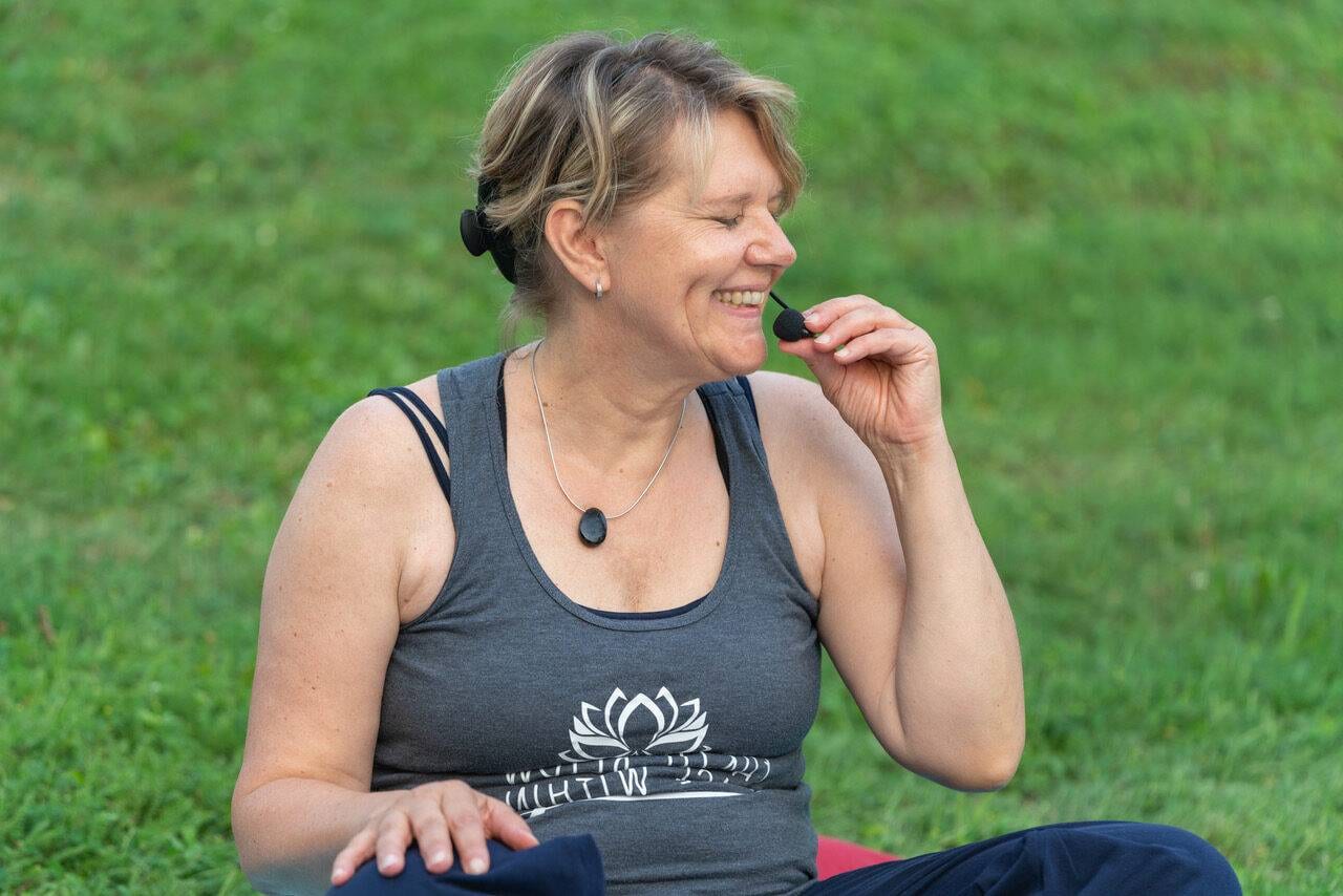 Manuela Förg sitzt im Gras und lacht, ein Headset-Mikrofon am Ohr