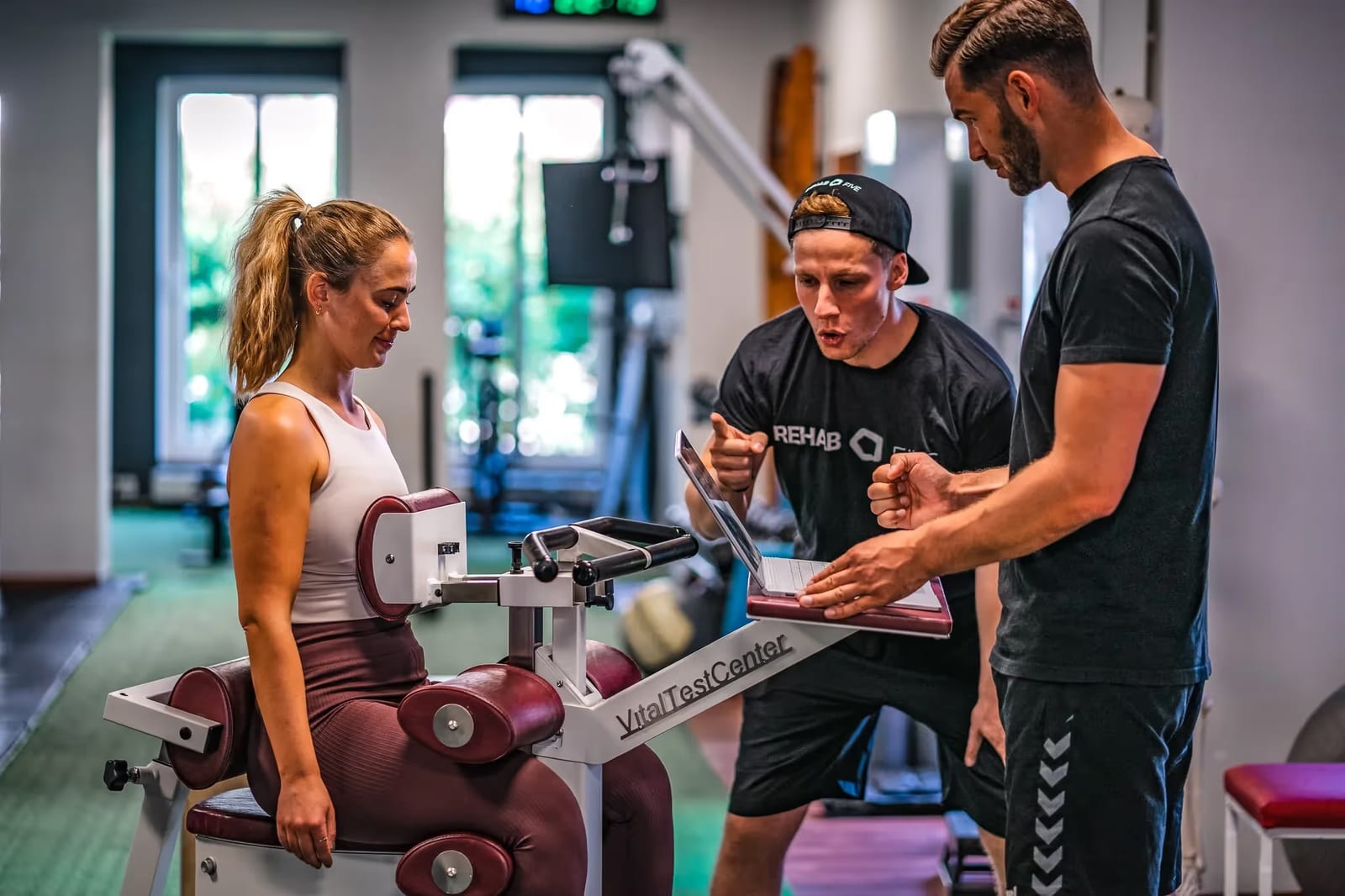 Ein Trainer von REHAB FIVE erklärt einer Teilnehmerin eine Übung an der Beinpresse, daneben schaut ein Kollege zu