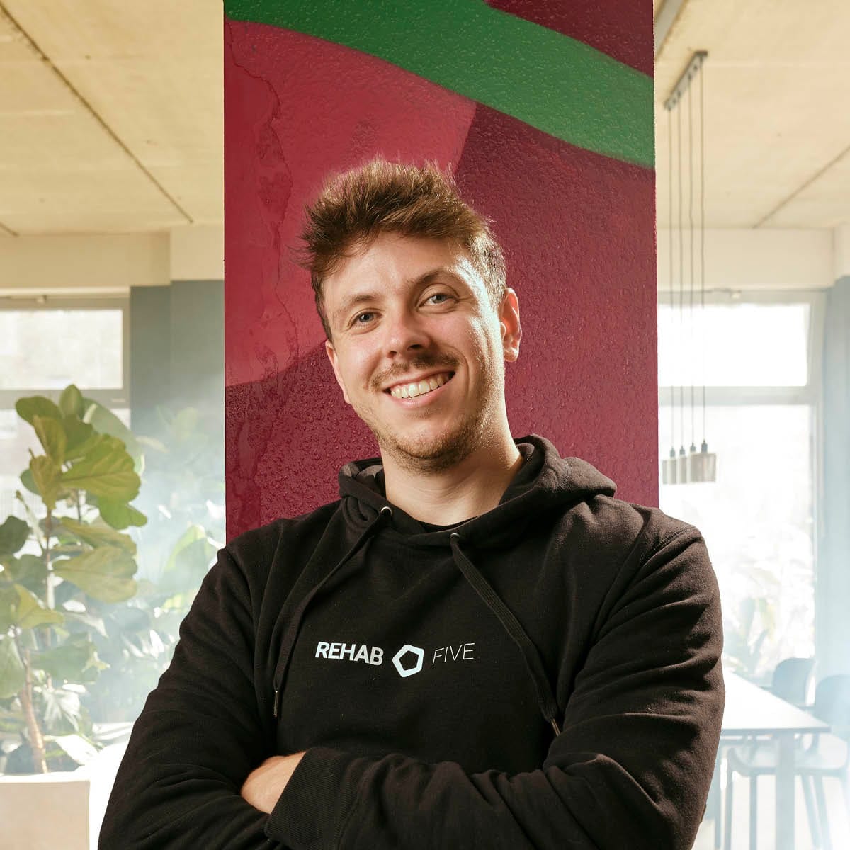 Niklas, Physiotherapeut bei REHAB FIVE, im schwarzen Teamhoodie mit verschränkten Armen