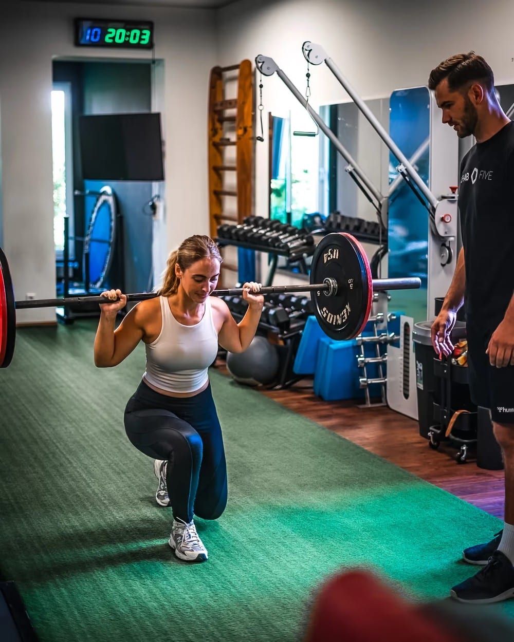 Eine Teilnehmerin macht eine Kniebeuge mit Langhantel, ein Trainer von REHAB FIVE steht daneben und korrigiert
