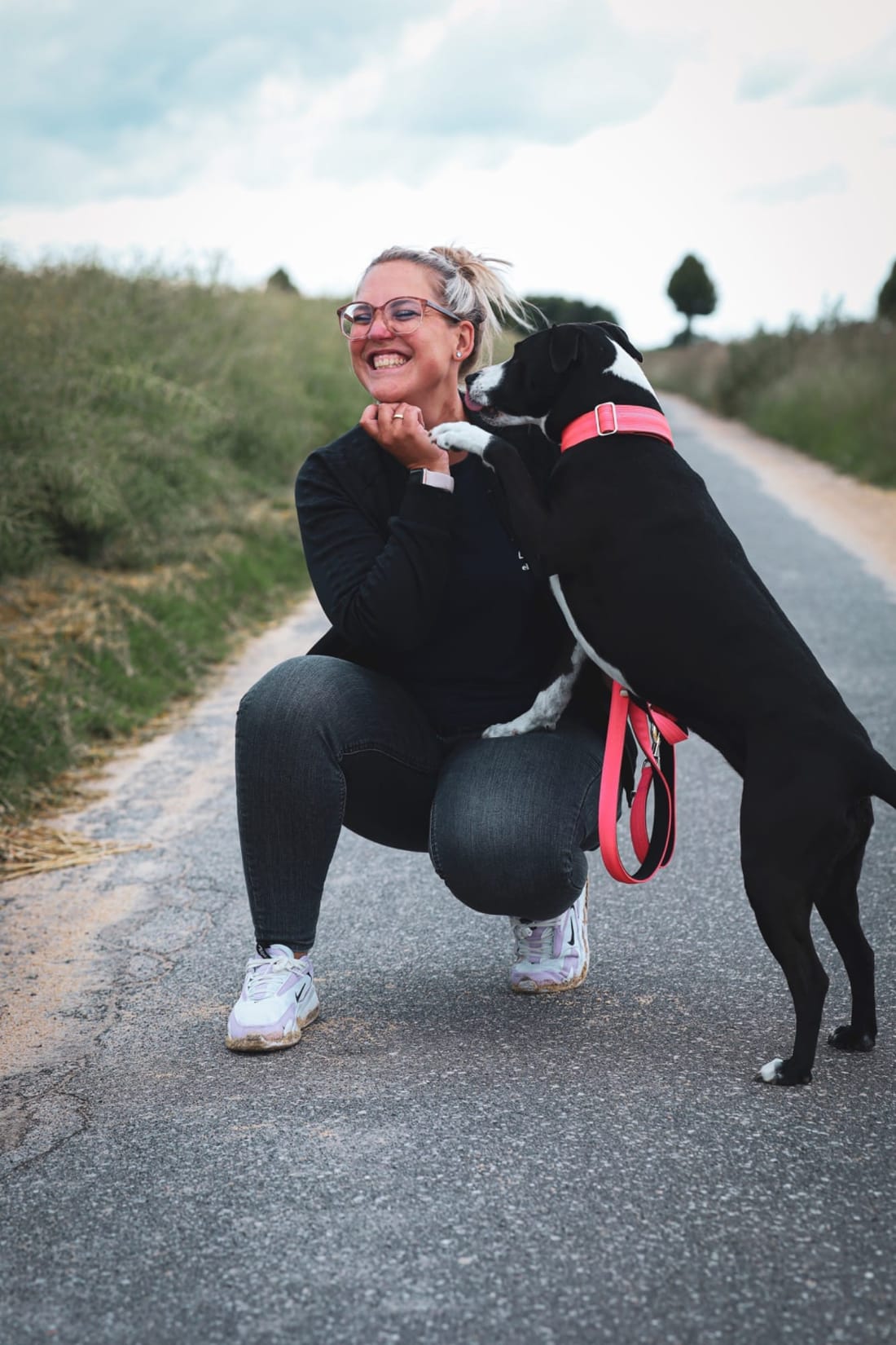 Maureen Smith kniet lachend auf einem Feldweg, ihr schwarz-weißer Hund an der pinken Leine legt ihr die Pfote auf die Schulter