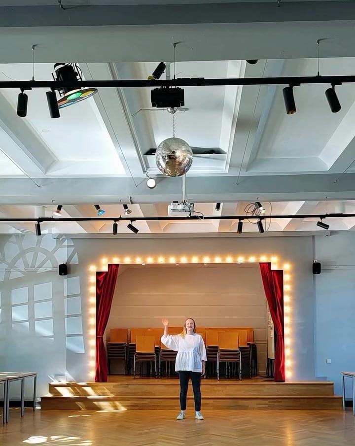 Lilian winkt im leeren Steenkampsaal, hinter ihr die Bühne mit Glühbirnenrahmen und roten Vorhängen, über ihr eine Discokugel