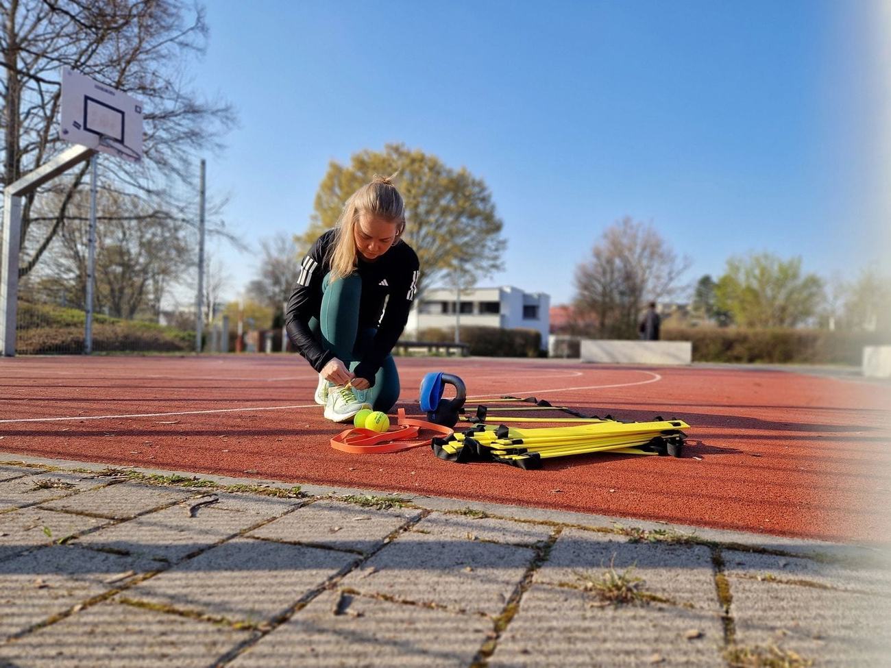 Sabrina schnürt die Schuhe neben Koordinationsleiter, Widerstandsband und Kettlebell auf der Tartanbahn